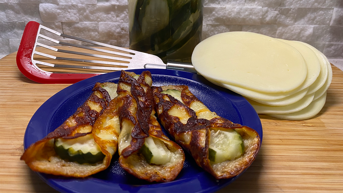 Three pickles-in-a-blanket served on a blue plate next to a jar of pickles, a stack of provolone cheese and a red and silver spatula