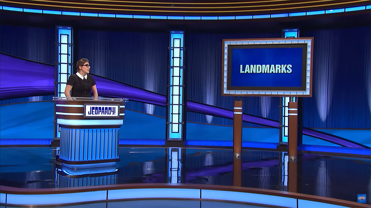 Host of "Jeopardy!" Mayim Bialik in a black shirt with white collar presents the final category on stage 'Landmarks'