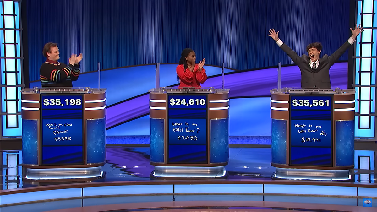 Contestants of "Jeopardy" Jackson in a black sweater with rainbow detail around the sleeves, Maya in a red blouse, and Justin in a suit with his arms up in the air, learn Jackson is the winner