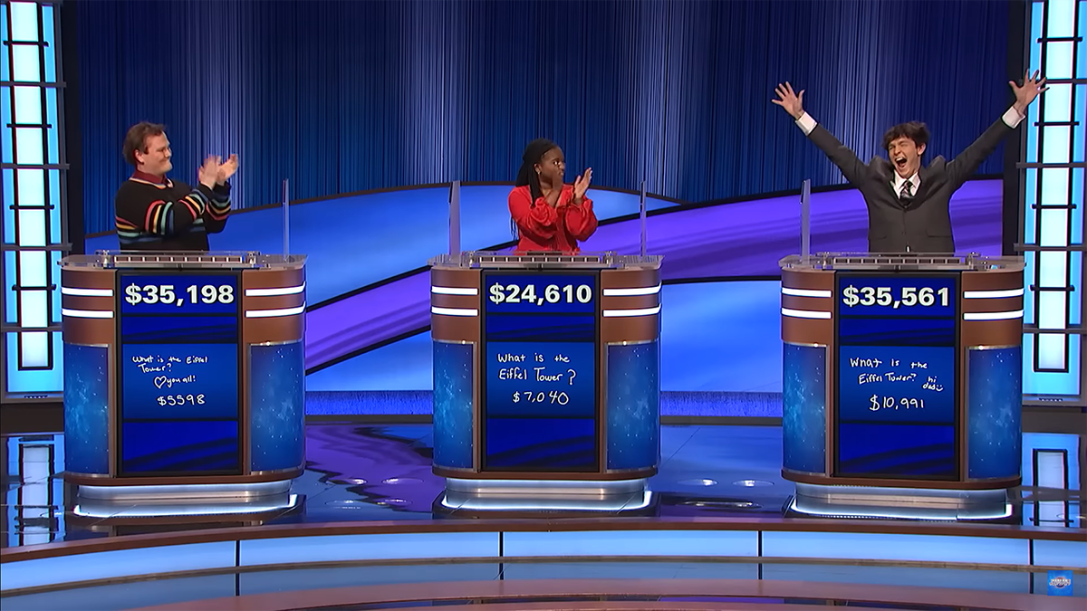 Contestants of "Jeopardy" Jackson in a black sweater with rainbow detail around the sleeves, Maya in a red blouse, and Justin in a suit with his arms up in the air, learn Jackson is the winner