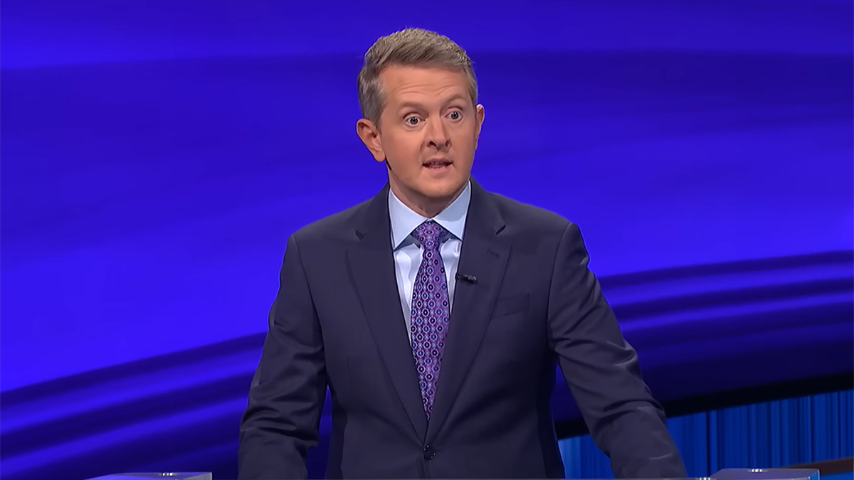 Ken Jennings in a navy suit with a light blue shirt and purple suit on the set of Jeopardy!"