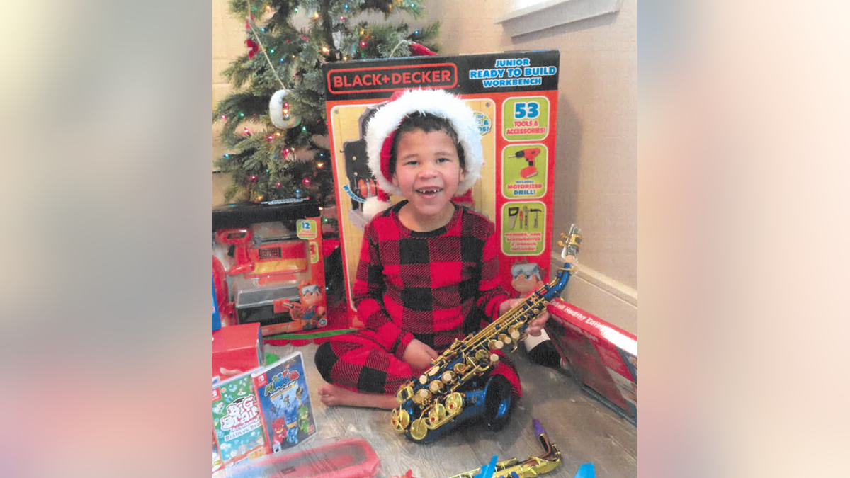 Zohar smiles in plaid (red and black) pajamas surrounded by toys and wearing a Santa hat, holding a toy saxaphone