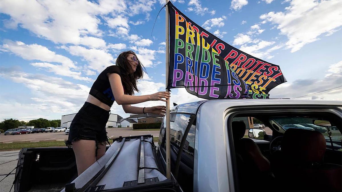 Woman standing in the back of a pickup truck holding a pride protest flag.