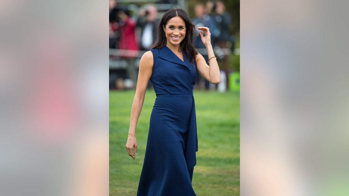 Meghan Markle wearing a sleeveless blue dress walking on grass and smiling
