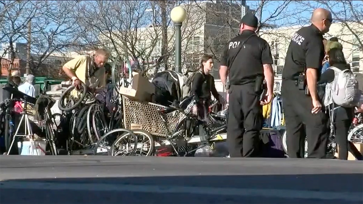 Police officers stand on a sidewalk strewn with bicycles, shopping carts and other items associated with a homeless camp in Denver