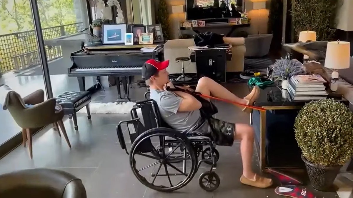 Jeremy Renner in a red hat and light blue/grey shirt sits in a wheelchair doing leg exercises in his home