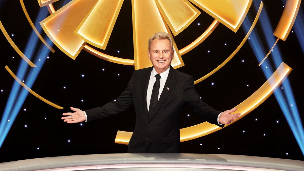 Pat Sajak proudly stands with arms wide open on "Wheel of Fortune" set.