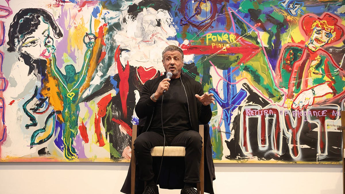 Sylvester Stallone talking in front of one of his paintings