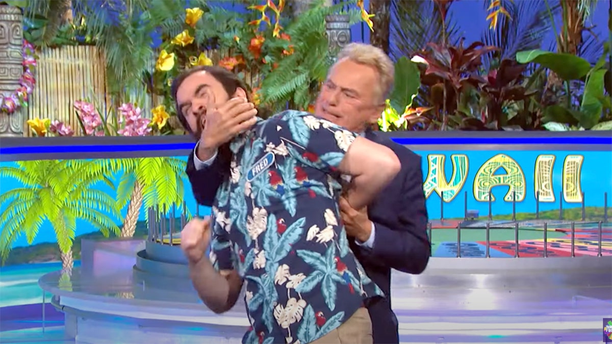 Contestant Fred in a patterned blue shirt has his arm locked behind him and neck locked by Pat Sajak as he jokingly is tackled
