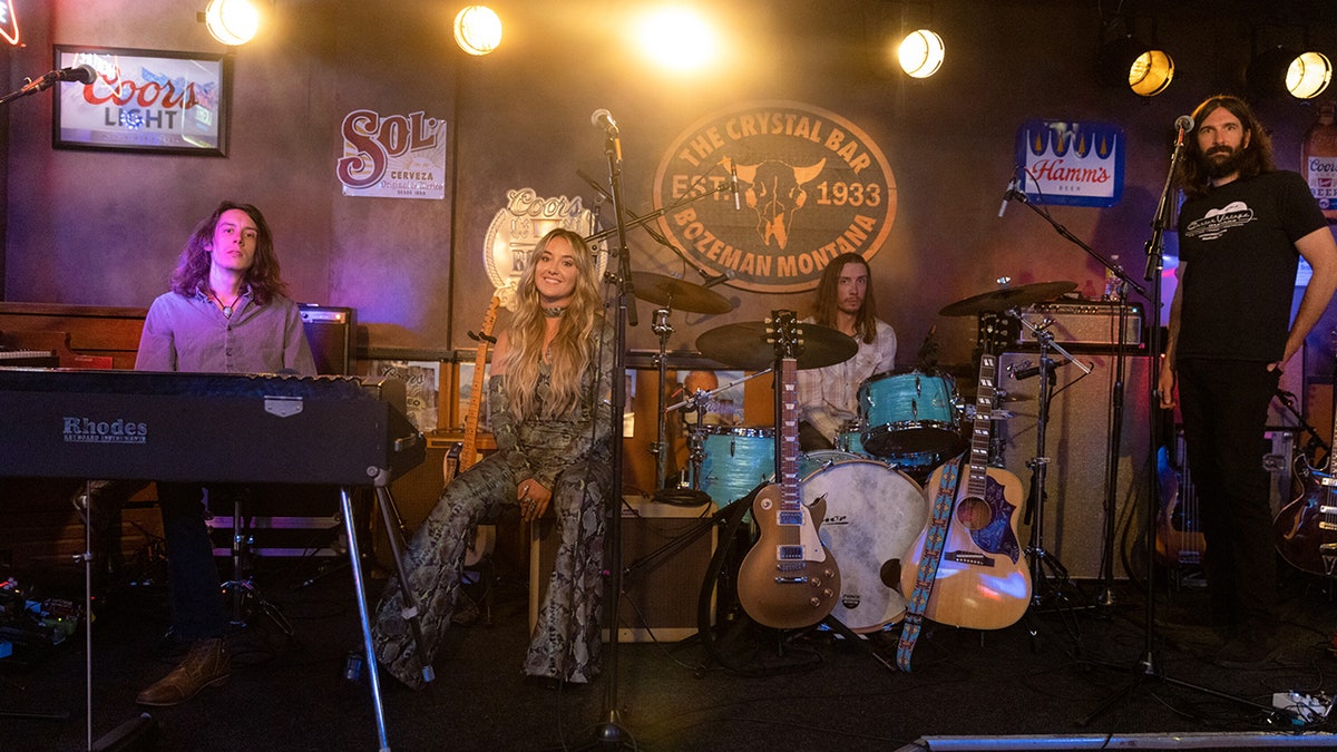 Lainey Wilson as Abby on "Yellowstone" in a black snakeskin jumpsuit sitting on stage smiling at the camera alongside her band mates