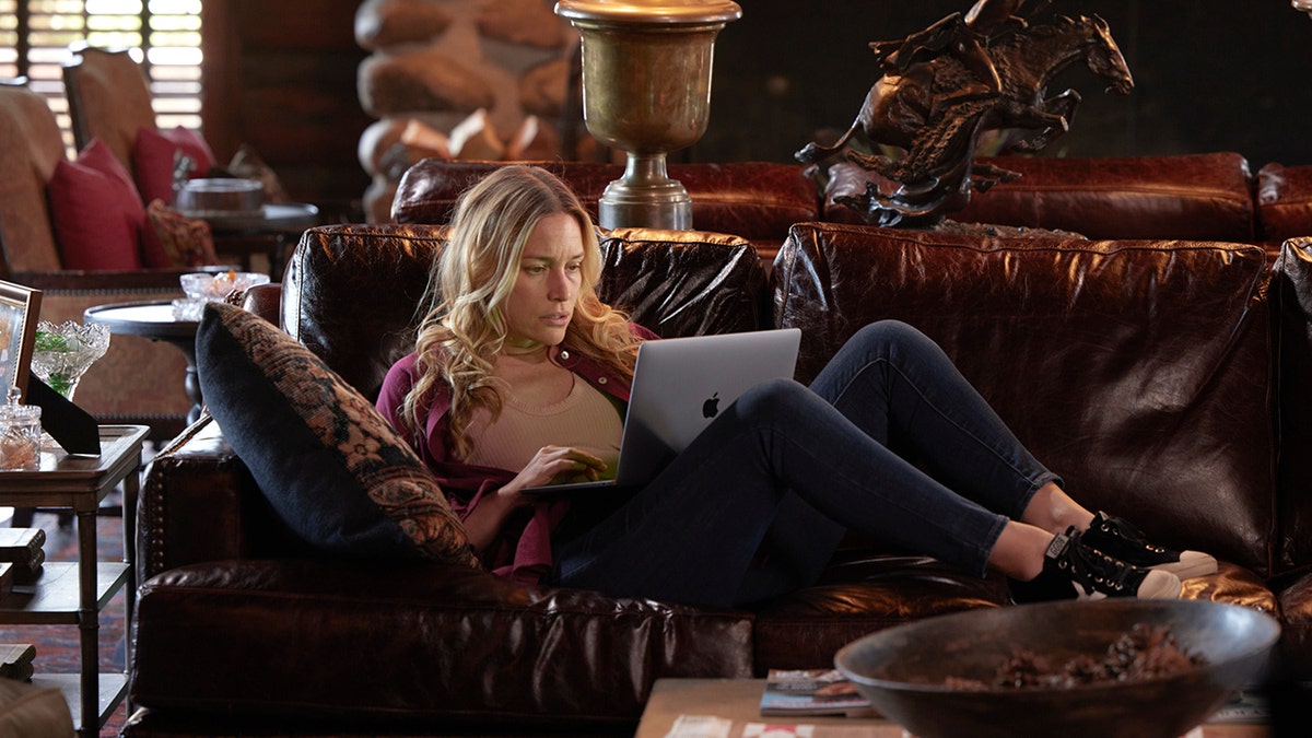 Piper Perabo as Summer Higgins on "Yelllowstone" lying on a brown leather couch in a red cardigan and jeans, staring intently at her Mac laptop 