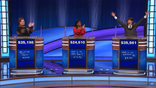 Contestants of "Jeopardy" Jackson in a black sweater with rainbow detail around the sleeves, Maya in a red blouse, and Justin in a suit with his arms up in the air, learn Jackson is the winner