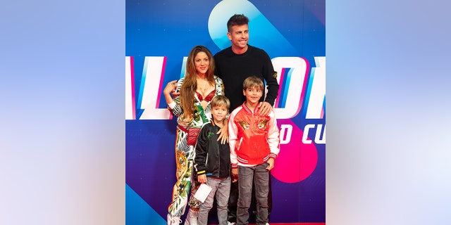 Shakira exposes her red leopard bra wearing a white outfit with jungle animals on it, has Gerard Piqué's arm wrapped around her, he is wearing black, and their two sons both in relaxed bomber jackets stand in front of them