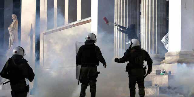 Seorang pengunjuk rasa berkerudung dengan masker gas air mata melemparkan alat pemadam api ke arah polisi anti huru hara selama bentrokan di markas Universitas Athena, di Yunani, pada 8 Maret 2023. Protes pecah terkait kecelakaan kereta api mematikan di Yunani yang menewaskan 57 orang. Tiga orang lagi didakwa sehubungan dengan kecelakaan kereta yang mematikan itu.
