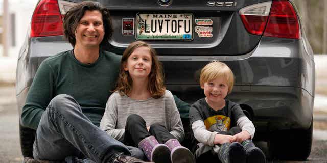 A man sits with his kids behind their car with a vanity license plate that the state of Maine has deemed in appropriate on March 8, 2023, in Poland, Maine. The vegan family's car will soon have a randomly selected plate.