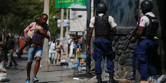 A parent, carrying his child after picking him up from school, runs past police carrying out an operation against gangs in the Bel-Air area of Port-au-Prince, Haiti, March 3, 2023. An American couple is reportedly missing in the same area.