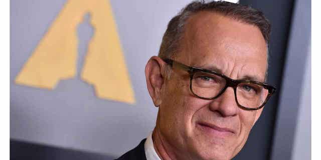Tom Hanks soft smiles on the red carpet in a dark suit and dark framed glasses