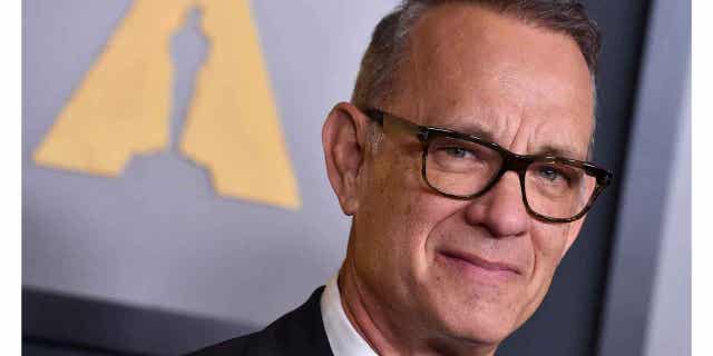 Tom Hanks soft smiles on the red carpet in a dark suit and dark framed glasses