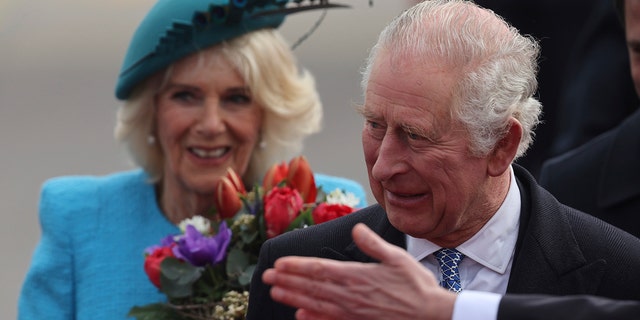 King Charles looks off to supporters as he arrives in Germany and a hand ushers him to the right, Camilla in a turquoise/teal outfit walks behind with a bouquet of flowers