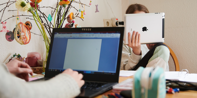 A mother works at the kitchen table while her daughter looks into a tablet across from her. Photo: Annette Riedl/dpa (Photo by Annette Riedl/picture alliance via Getty Images)