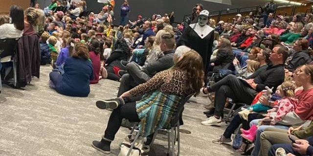 A drag queen protester dressed in black and white is shown moving throughout the crowd during the book reading event in Fayetteville, Arkansas. Protesters are also shown holding signs along the far wall.