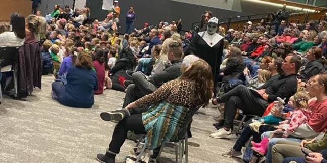 A drag queen protester dressed in black and white is shown moving throughout the crowd during the book reading event in Fayetteville, Arkansas. Protesters are also shown holding signs along the far wall.