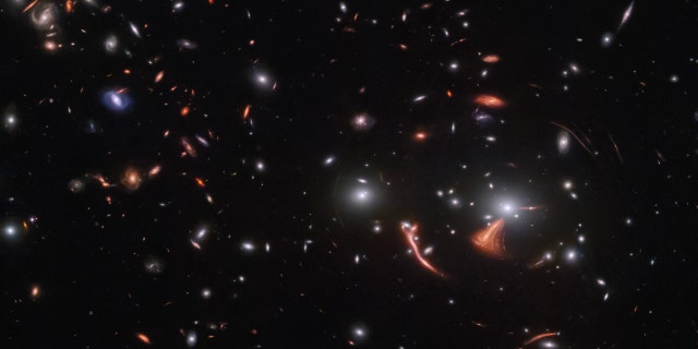 Many small galaxies are scattered on a black background: mainly, white, oval-shaped and red, spiral galaxies. To the lower right is a galaxy cluster, with a very large and bright elliptical galaxy at its center. Thin, reddish, stretched-out arcs surround it. One arc is thick and much brighter. Another red galaxy is large and warped, just next to the cluster core.