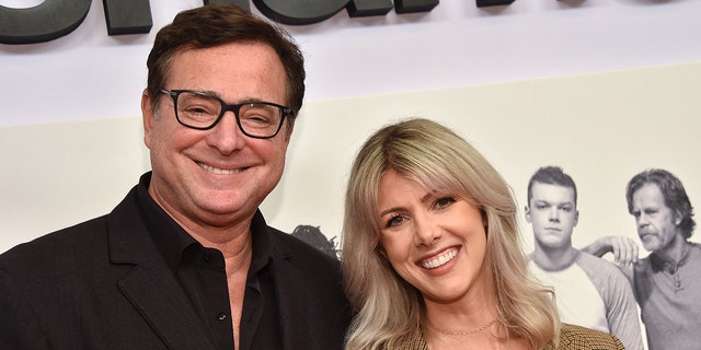 Bob Saget in a black shirt with black frames smiles next to wife Kelly Rizzo in a tan jacket on the red carpet