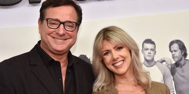 Bob Saget in a black shirt with black frames smiles next to wife Kelly Rizzo in a tan jacket on the red carpet