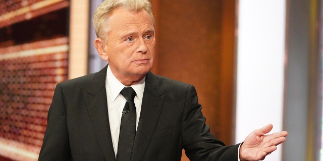 Pat Sajak in a black suit, white button down, and black tie looks off-camera while filming "Celebrity Wheel of Fortune"