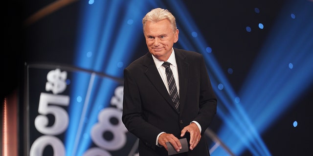 Pat Sajak stands behinds a barrier wearing a black suit and a patterned tie holding a card taping "Celebrity Wheel of Fortune"