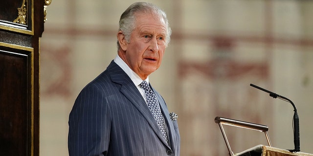 King Charles at the podium wearing a blue pin-stripe suit speaking in Westminster Abbey for Commonwealth Day
