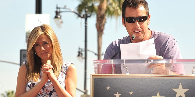 Adam Sandler giving a speech at Jennifer Aniston's Walk of Fame ceremony, with her standing behind him.