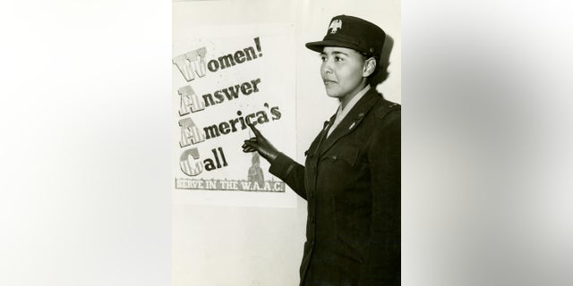 African American Charity Adams, First Officer in the Women's Army Auxiliary Corps, standing in uniform and pointing to a poster that reads, 'Women! Answer America's Call, Serve in the W.A.A.C.,' 1943-02.