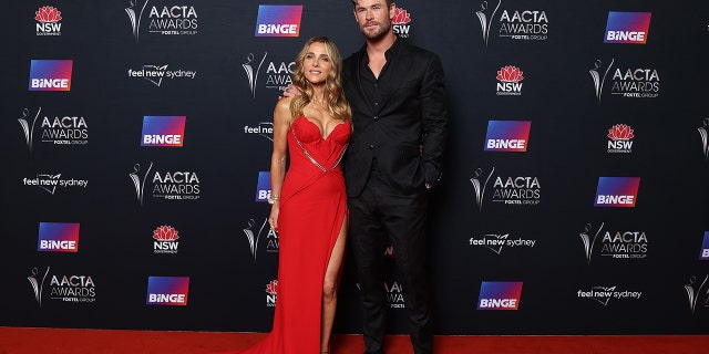 Chris Hemsworth in a black suit and jacket poses on the red carpet next to Elsa Pataky in a busty red gown in Sydney, Australia