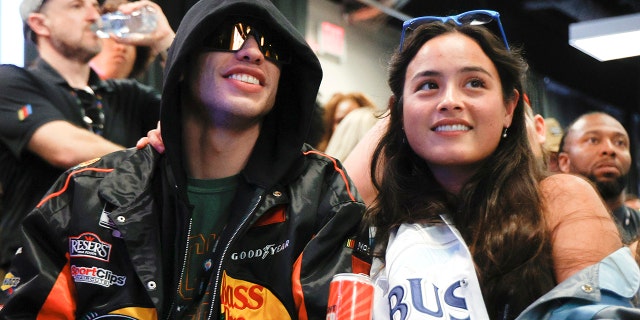 Pete Davidson in a black sweatshirt with his hood up and sunglasses on smiles next to Chase Sui Wonders at the Daytona 500 race in Florida