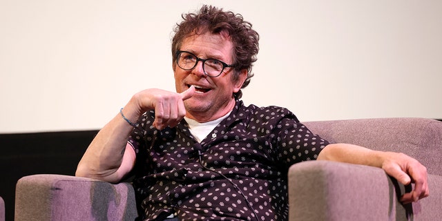 Майкл Дж. Фокс сидит в загорелом кресле на сцене SXSW, в черном топе с белыми цветами и джинсах, в очках в черной оправе.