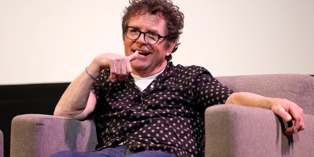 Michael J. Fox sits in a tan chair on the SXSW stage, in a black top with white flowers and jeans, wearing black rimmed glasses