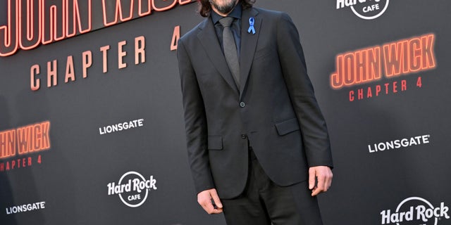 Keanu Reeves wearing a suit with a blue ribbon pinned to his lapel
