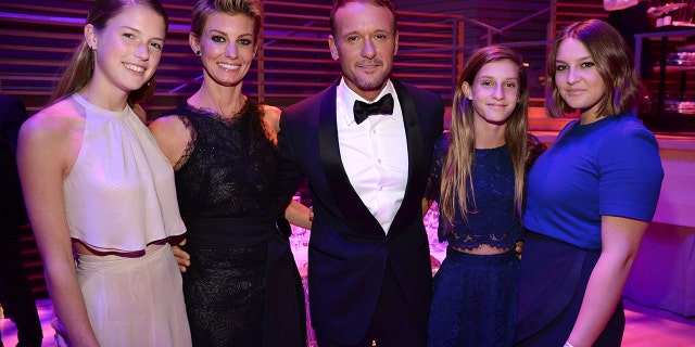 Maggie McGraw in a blush halter top and skirt smiles next to mother Faith Hill in a long black dress, husband Tim McGraw in a classic black tuxedo, daughter Audrey in a blue dress, and daughter Gracie in a blue dress