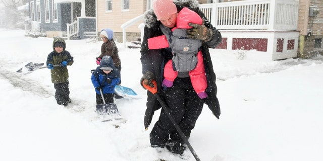 Desiree Besner clears the sidewalk of snow with her youngest child, Camille, strapped to her chest while enlisting the help of her other children, Ryu, from left, Ezra, and Karma at their home in Auburn, N.Y., Tuesday, March 14, 2022. 