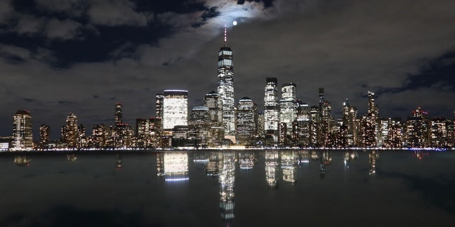 La pleine lune de ver se lève par une soirée nuageuse derrière l'horizon du bas Manhattan et du One World Trade Center à New York, alors qu'elle se reflète sur le sommet d'un monument le 7 mars 2023, vue de Jersey City, New Jersey. 