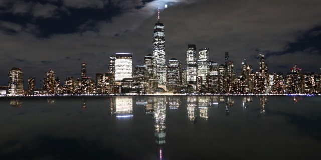 La pleine lune de ver se lève par une soirée nuageuse derrière l'horizon du bas Manhattan et du One World Trade Center à New York, alors qu'elle se reflète sur le sommet d'un monument le 7 mars 2023, vue de Jersey City, New Jersey.