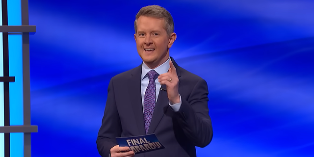 Ken Jennings in a navy suit with a light blue shirt and purple suit on the set of 'Jeopardy!' pointing at the camera