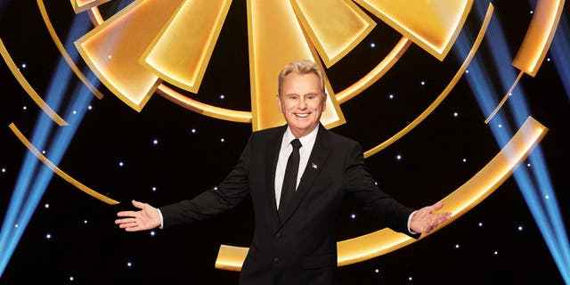 Pat Sajak proudly stands with arms wide open on "Wheel of Fortune" set.