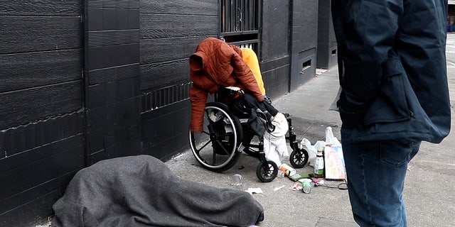 Kevin Dahlgren approaches a man in a wheelchair who is missing both his legs. A woman wrapped in a blanket lies on the sidewalk nearby.