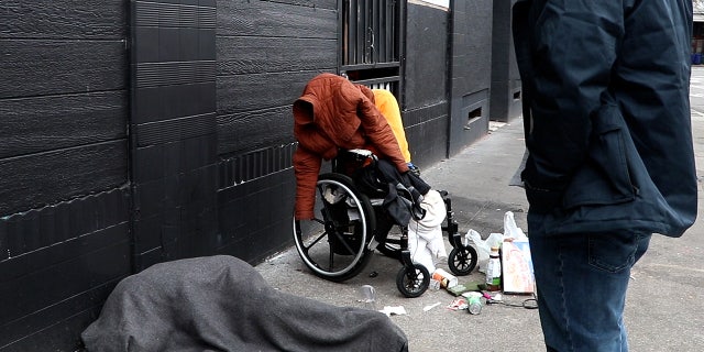 Kevin Dahlgren approaches a man in a wheelchair who is missing both his legs. A woman wrapped in a blanket lies on the sidewalk nearby.