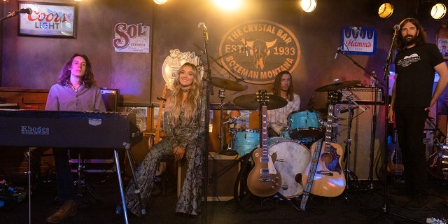 Lainey Wilson as Abby on "Yellowstone" in a black snakeskin jumpsuit sitting on stage smiling at the camera alongside her band mates