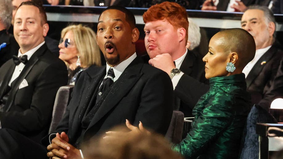 Will Smith in a black suit sits next to his wife Jada Pinkett Smith in a long cascading green gown with his mouth open at the Oscars