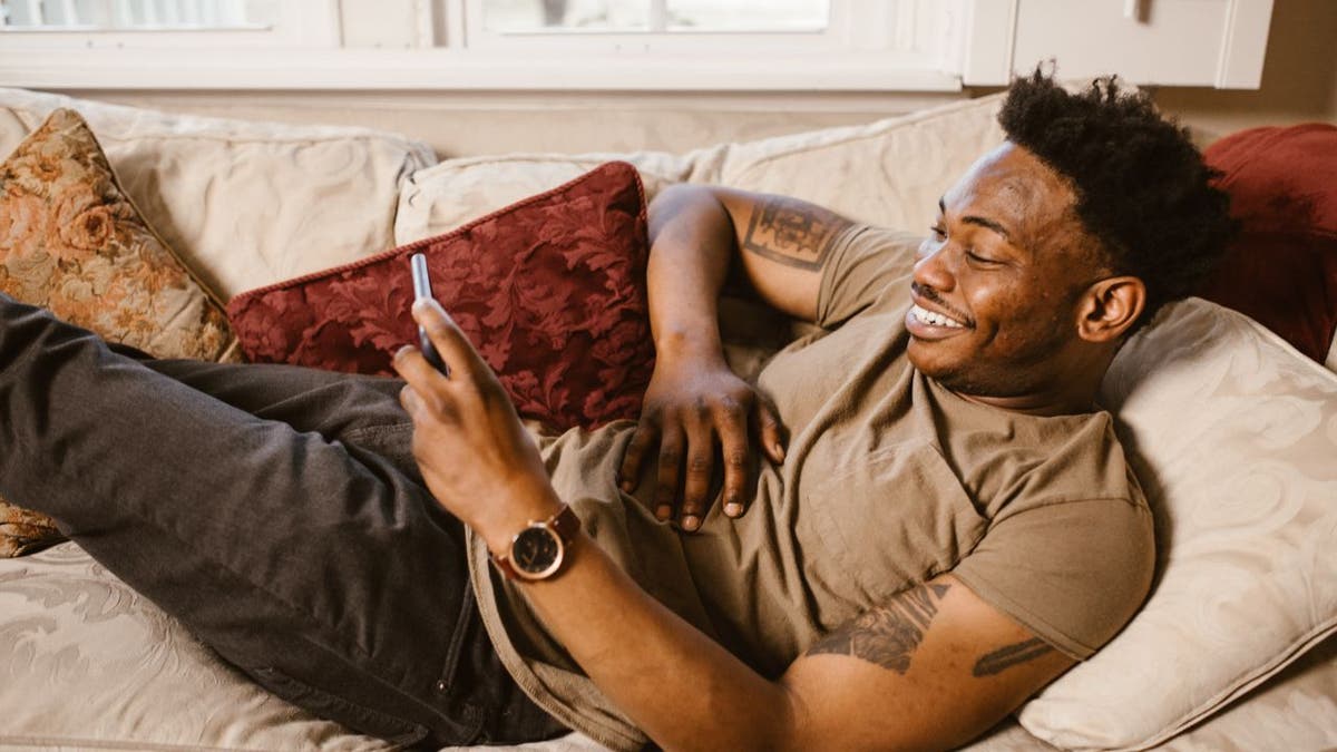 Man laying on his couch while smiling at his phone.